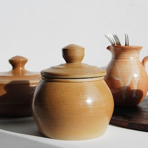 Peut inclure: Ensemble de poterie artisanale de couleur beige. L'ensemble comprend des pots à couvercle, un petit pichet avec une anse et un vase plus grand. Des fourchettes sont placées dans le pichet. Les articles sont disposés sur un plateau en bois.