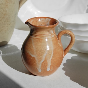 Op de afbeelding: Een kleine, ronde keramische kan met een handvat en tuit. De kan heeft een warme, aardse oranje kleur met witte strepen en een getextureerd oppervlak. Het is omgeven door witte kommen en een grotere beige kan.