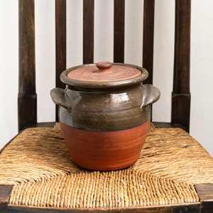 May include: A brown and orange ceramic pot with a lid sits on a woven chair seat.