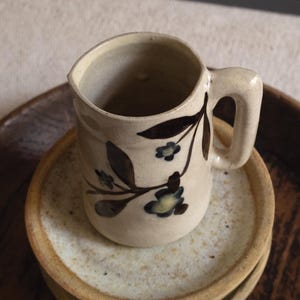 Könnte beinhalten: Eine kleine, cremefarbene Keramiktasse mit Henkel, verziert mit braunen Blumenranken und blauen Blüten. Die Tasse steht auf einem Stapel aus zwei Keramiktellern und einer Holzschale.