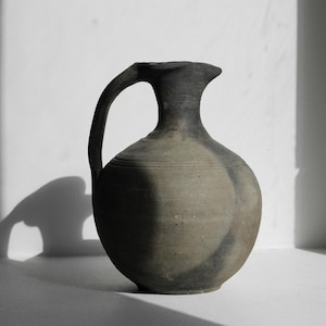 May include: A dark grey ceramic jug with a curved handle and a rounded body. The jug has a textured surface and a flared rim. The pitcher is displayed against a white background, with shadows adding depth. The jug is likely handmade.