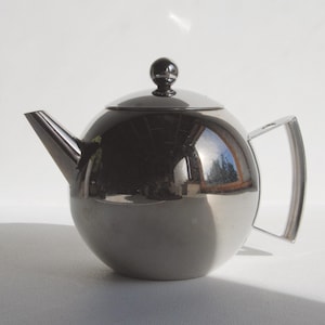 Op de afbeelding: Een glanzende, zilverkleurige theepot met een ronde body, een tuit en een handvat. Het deksel heeft een kleine, ronde knop. De theepot reflecteert de omgeving, waardoor een spiegeleffect ontstaat. De theepot is ontworpen voor het zetten van thee.
