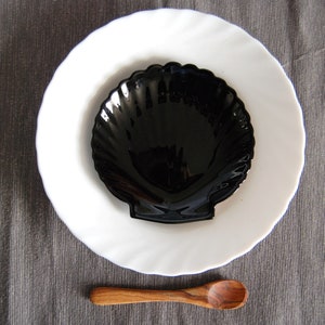May include: A black, shell-shaped dish sits on a white, scalloped-edge plate. A small, wooden spoon rests below. The items are arranged on a grey, textured surface, creating a simple, elegant still life.