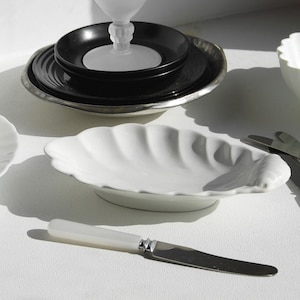 May include: A selection of white and black serving dishes and utensils. Includes a white shell-shaped dish, a black plate, a silver-rimmed plate, and a white bowl. Also, a butter knife and serving utensils with white handles.