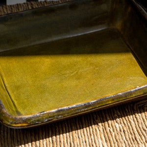 May include: A rectangular ceramic baking dish with a dark green glaze. The dish is empty and sitting on a woven surface.