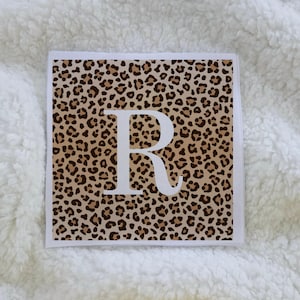 May include: Square decorative pillow with a leopard print pattern and a large white letter "R" in the center. The pillow has a white border and is set against a white, textured background.