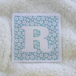 May include: A square, white card featuring a large, white letter "R". The background of the card is a light blue color with a floral pattern. The card is resting on a white, fluffy surface.
