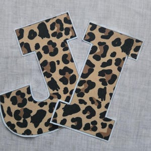 May include: Two fabric letters, "J" and "V", are displayed. The letters have a tan background with black and brown leopard spots. Each letter is outlined in a light blue stitch.