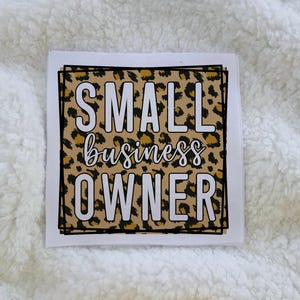 May include: Square sign with the words "SMALL business OWNER" in white block letters and cursive on a leopard print background, framed by a black border. The sign is on a white background.