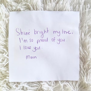 May include: A white square note with purple handwritten text that reads "Shine bright my love, I'm so proud of you. I love you. Mom." The note is on a fluffy white surface.