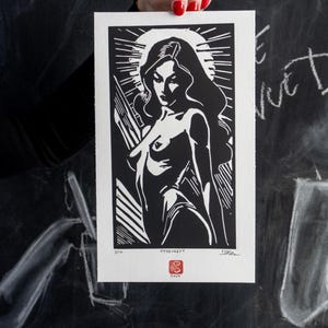 May include: Black and white linocut print of a woman with long hair, set against a radiant sunburst. The artwork is held up, showcasing the print's details and the artist's signature. The print has a red stamp.
