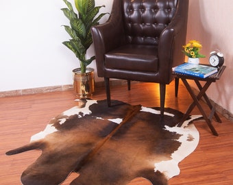 Puede incluir: Un sillón de cuero marrón con tapicería de botones se encuentra sobre una alfombra de piel de vaca marrón y blanca. Una pequeña mesa de madera con un libro y un reloj se encuentra junto al sillón.