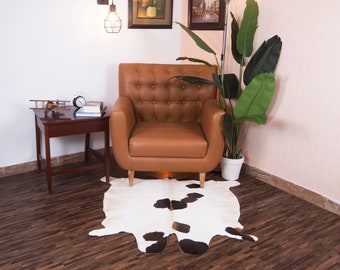 Puede incluir: Un sillón de cuero marrón con tapicería de botones se encuentra sobre una alfombra de piel de vaca en una sala de estar con suelo de madera. En el fondo hay una planta en maceta y una mesa auxiliar con un reloj y libros.