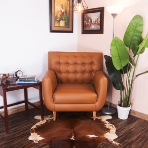 Puede incluir: Un sill&oacute;n de cuero marr&oacute;n con tapicer&iacute;a de botones se encuentra sobre una alfombra de piel de vaca marr&oacute;n y blanca en una sala de estar. El sill&oacute;n est&aacute; frente a una pared blanca con dos cuadros enmarcados y una l&aacute;mpara colgante. Una planta en maceta con grandes hojas verdes se encuentra en la esquina de la habitaci&oacute;n.