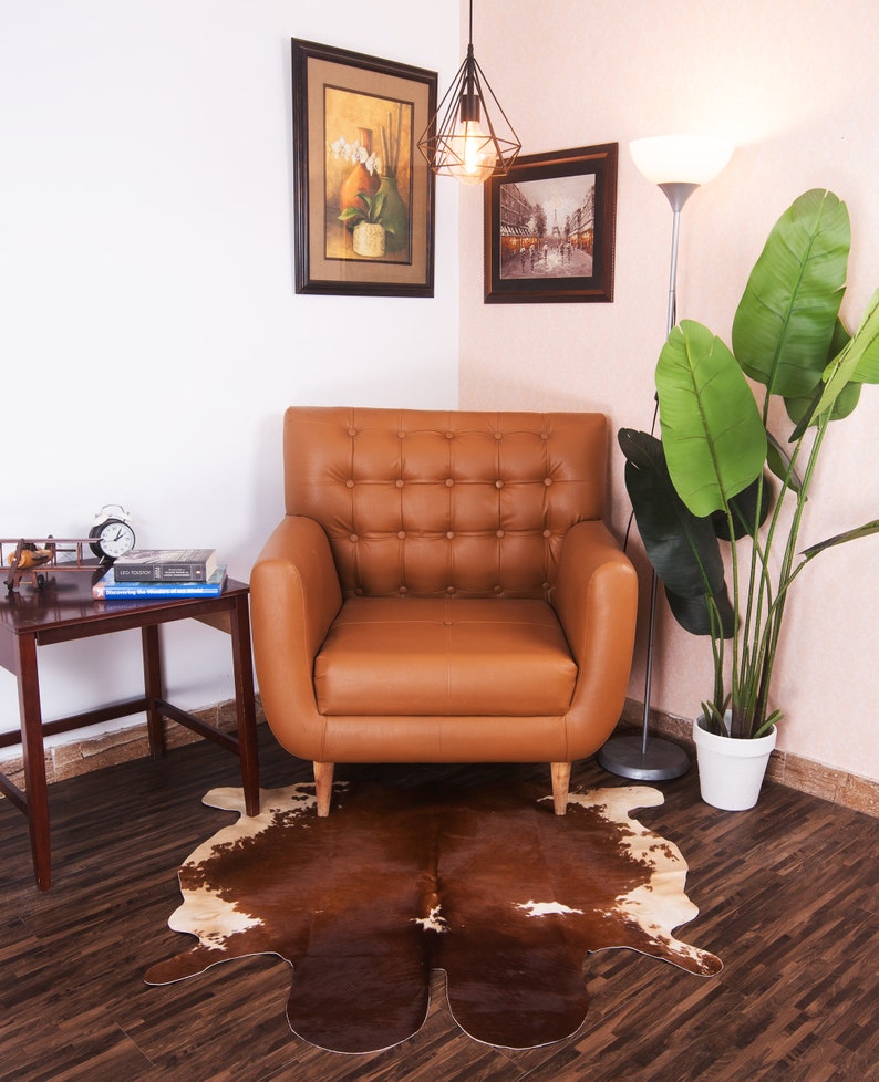 Puede incluir: Un sill&oacute;n de cuero marr&oacute;n con tapicer&iacute;a de botones se encuentra sobre una alfombra de piel de vaca marr&oacute;n y blanca en una sala de estar. El sill&oacute;n est&aacute; frente a una pared blanca con dos cuadros enmarcados y una l&aacute;mpara colgante. Una planta en maceta con grandes hojas verdes se encuentra en la esquina de la habitaci&oacute;n.