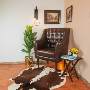 Puede incluir: Un sill&oacute;n de cuero marr&oacute;n con tapicer&iacute;a de botones se encuentra sobre una alfombra de piel de vaca marr&oacute;n y blanca en una habitaci&oacute;n con suelo de madera. Una peque&ntilde;a mesa de madera con un libro y un reloj se encuentra junto al sill&oacute;n. Una planta en maceta y una l&aacute;mpara de pared con una bombilla se encuentran en el fondo.