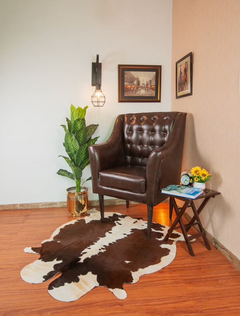 Puede incluir: Un sill&oacute;n de cuero marr&oacute;n con tapicer&iacute;a de botones se encuentra sobre una alfombra de piel de vaca marr&oacute;n y blanca en una habitaci&oacute;n con suelo de madera. Una peque&ntilde;a mesa de madera con un libro y un reloj se encuentra junto al sill&oacute;n. Una planta en maceta y una l&aacute;mpara de pared con una bombilla se encuentran en el fondo.