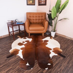 Puede incluir: Una alfombra de piel de vaca marr&oacute;n y blanca con una forma natural irregular. La alfombra est&aacute; colocada sobre un suelo de madera dura en un sal&oacute;n con un sill&oacute;n de cuero marr&oacute;n y una mesa auxiliar.