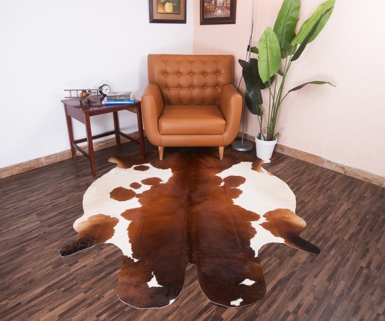 Puede incluir: Una alfombra de piel de vaca marr&oacute;n y blanca con una forma natural irregular. La alfombra est&aacute; colocada sobre un suelo de madera dura en un sal&oacute;n con un sill&oacute;n de cuero marr&oacute;n y una mesa auxiliar.