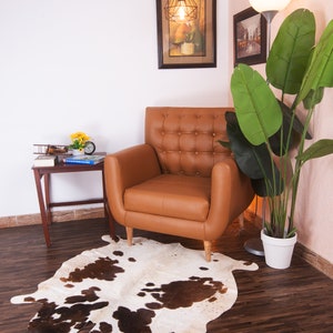 Puede incluir: Un sill&oacute;n de cuero marr&oacute;n con tapicer&iacute;a de botones se encuentra sobre una alfombra de piel de vaca blanca y marr&oacute;n en una sala de estar. Una planta en maceta con grandes hojas verdes est&aacute; en la esquina, y una peque&ntilde;a mesa auxiliar de madera con un libro y un avi&oacute;n de juguete est&aacute; al lado del sill&oacute;n.