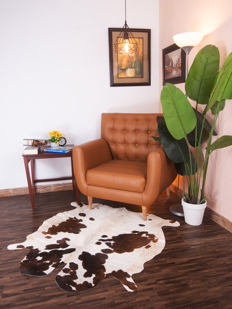 Puede incluir: Un sill&oacute;n de cuero marr&oacute;n con tapicer&iacute;a de botones se encuentra sobre una alfombra de piel de vaca blanca y marr&oacute;n en una sala de estar. Una planta en maceta con grandes hojas verdes est&aacute; en la esquina, y una peque&ntilde;a mesa auxiliar de madera con un libro y un avi&oacute;n de juguete est&aacute; al lado del sill&oacute;n.
