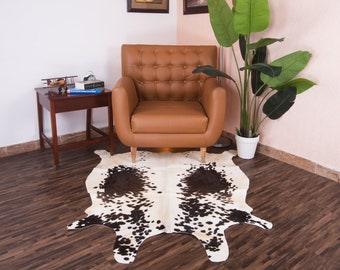 Puede incluir: Un sillón de cuero marrón con tapicería de botones se encuentra sobre una alfombra de piel de vaca en blanco y negro en un ambiente de sala de estar. Una pequeña mesa auxiliar de madera con libros y un avión de modelo se encuentra junto al sillón. Una planta verde en maceta con hojas grandes se encuentra en la esquina.