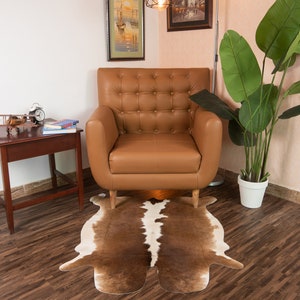 Puede incluir: Un sill&oacute;n de cuero marr&oacute;n con tapicer&iacute;a de botones se encuentra sobre una alfombra de piel de vaca marr&oacute;n y blanca. Una peque&ntilde;a mesa auxiliar de madera con un avi&oacute;n de modelo y libros se encuentra junto al sill&oacute;n.