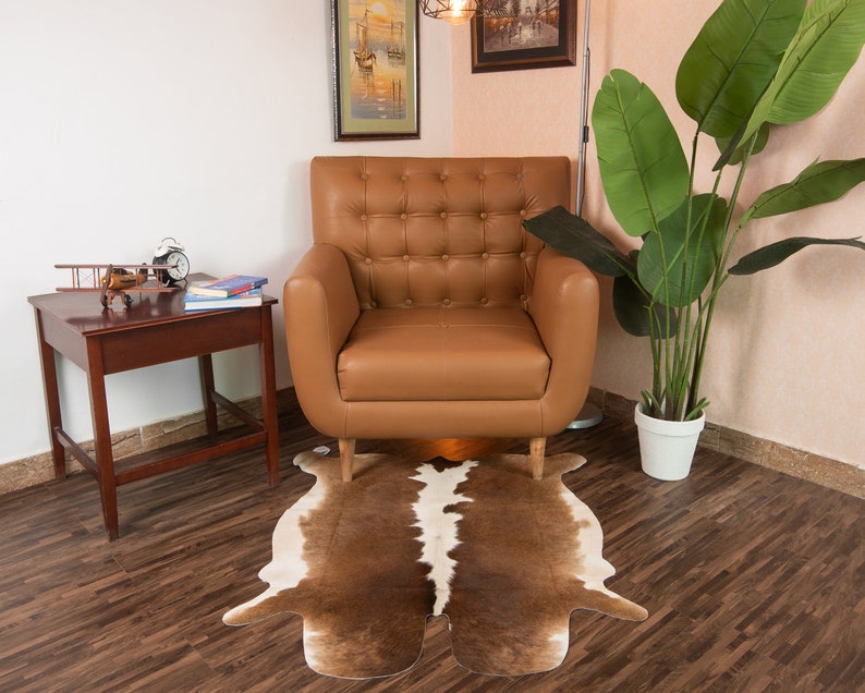 Puede incluir: Un sill&oacute;n de cuero marr&oacute;n con tapicer&iacute;a de botones se encuentra sobre una alfombra de piel de vaca marr&oacute;n y blanca. Una peque&ntilde;a mesa auxiliar de madera con un avi&oacute;n de modelo y libros se encuentra junto al sill&oacute;n.