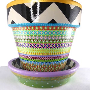 May include: A hand-painted terracotta planter with a chevron pattern in black and white on the top and a colorful geometric pattern on the bottom. The planter has a matching saucer with a green base and a purple rim.