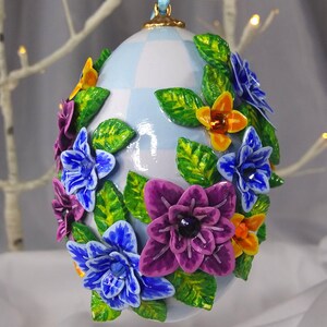 Könnte beinhalten: Ein blaues und weißes kariertes Osterei-Ornament, verziert mit lila, blauen und gelben Blumen und grünen Blättern.