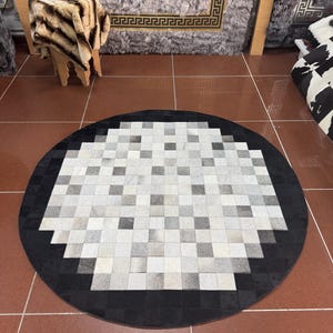 Peut inclure: Un tapis circulaire composé de carrés individuels en peau de vache. Le centre présente une grille de carrés blancs, gris clair et gris foncé, tandis que la bordure extérieure est constituée de carrés noirs unis disposés en cercle.