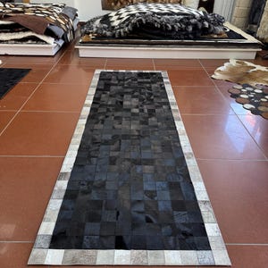 Peut inclure: Un tapis rectangulaire composé d'un assemblage de carrés de peau. La partie centrale présente une grille de carrés noirs unis, tandis que la bordure est constituée de carrés de peau plus clairs, aux tons gris et blancs mouchetés.