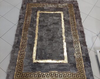 Puede incluir: Una alfombra rectangular hecha de piezas de piel gris texturizada en estilo patchwork. La alfombra presenta un borde dorado prominente con un patrón de greca negra en el centro del marco dorado, creando un diseño rectangular distintivo.