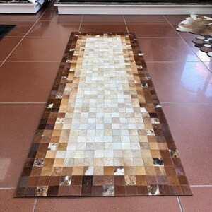 May include: A rectangular cowhide rug with a patchwork design. The rug has a mosaic pattern of squares in shades of brown, tan, and cream. The border is a darker brown, and the centre is lighter. The rug is displayed on a tiled floor.