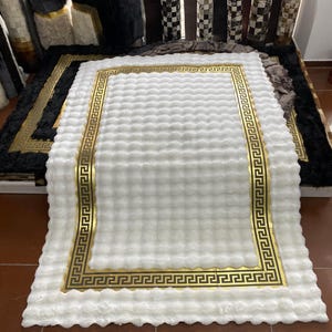 Op de afbeelding: Een witte, zachte vloerkleed met een gouden Griekse sleutelrand. Het vloerkleed is rechthoekig en heeft een zacht, getextureerd oppervlak. Andere vloerkleden in verschillende kleuren en patronen zijn zichtbaar op de achtergrond.