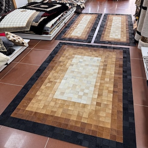 Puede incluir: Una exhibición de varias alfombras de piel de vaca en varios tamaños y diseños. Las alfombras presentan un patrón de patchwork con tonos de marrón, beige y negro. Algunas alfombras son rectangulares, mientras que otras están apiladas en una estantería. El suelo es de baldosas de color marrón rojizo.