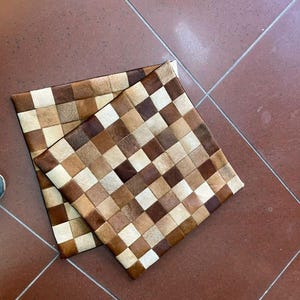 Puede incluir: Dos cojines cuadrados con un diseño de patchwork. Los cojines están hechos de cuero en tonos marrones, beige y crema, dispuestos en un patrón de cuadros. Los cojines están colocados sobre un suelo de baldosas marrones.