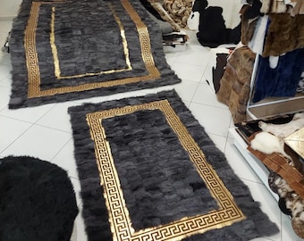 Puede incluir: Dos alfombras rectangulares de piel gris oscuro con un borde dorado prominente que presenta un patrón de greca repetitivo. Las alfombras tienen una textura de retazos y se exhiben sobre un suelo de baldosas blancas.