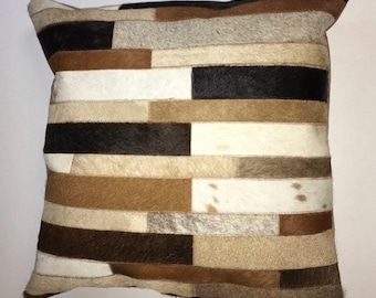 May include: Square decorative pillow with a patchwork design. The pillow features rectangular pieces of various colors, including black, white, brown, and tan. The pillow has a soft, textured appearance and is likely made of a natural material.