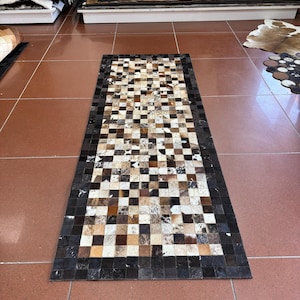 May include: A rectangular cowhide rug with a patchwork design. The rug features a checkerboard pattern in shades of brown, black, and white. The border is a solid black. The rug is displayed on a brown tile floor.