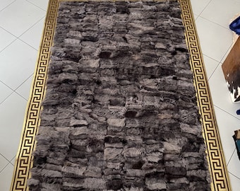 Puede incluir: Una alfombra rectangular con un panel central de piezas de piel texturizada de color gris oscuro en diseño patchwork. La alfombra está enmarcada por un borde ancho de color dorado decorado con un patrón de greca negra repetitivo, creando un contraste entre el centro y el borde.