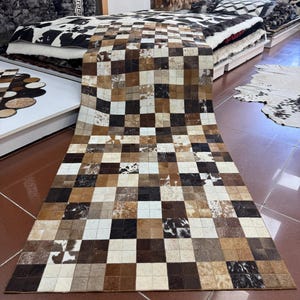 May include: A rectangular cowhide rug with a checkerboard pattern in shades of brown, tan, and white. The rug is made of individual squares of hide, creating a patchwork design. The rug is displayed in a store setting.