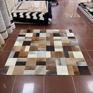 Puede incluir: Una alfombra cuadrada compuesta por parches rectangulares y cuadrados de piel de vaca en varios tonos de marrón, bronceado, blanco, gris y negro. Los parches están dispuestos en un patrón de cuadrícula geométrica en toda la superficie.