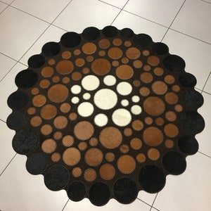 Op de afbeelding: Een rond, bruin, zwart en wit leren vloerkleed met een cirkelvormig patroon van verschillende grootte cirkels. Het vloerkleed is gemaakt van koeienhuid en heeft een gefranst randje.