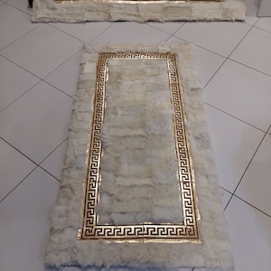May include: Two rectangular rugs made of off-white, textured fur patches. Each rug features a prominent, metallic gold border with a repeating Greek key pattern. The rugs are arranged with one positioned horizontally and one vertically.
