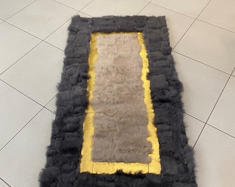 Puede incluir: Una alfombra rectangular con un borde grueso de pelo largo gris oscuro que rodea un panel central de piel beige claro. Un marco rectangular liso de color dorado metálico separa el borde gris oscuro del centro beige claro.