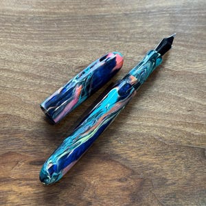 May include: A colorful fountain pen with a marbled design in shades of blue, teal, orange, and pink. The pen has a black nib and a matching cap, lying on a wooden surface. The pen is a writing instrument.