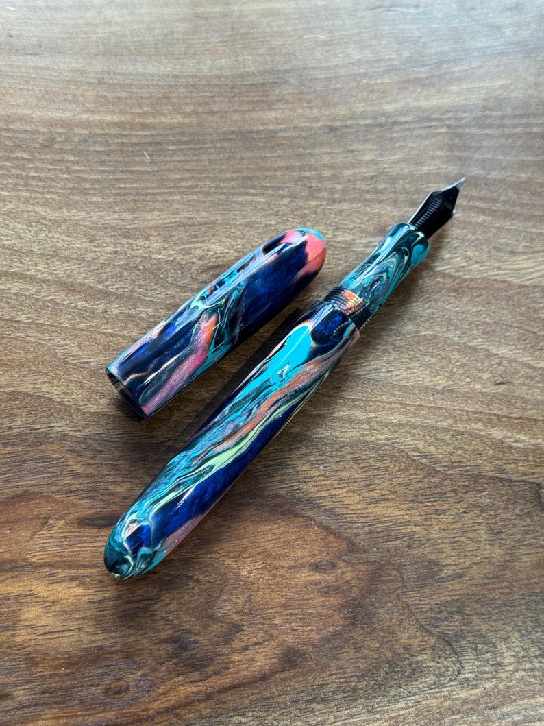 May include: A colorful fountain pen with a marbled design in shades of blue, teal, orange, and pink. The pen has a black nib and a matching cap, lying on a wooden surface. The pen is a writing instrument.