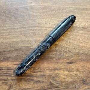 May include: A black and gray marbled fountain pen with a rounded tip and cap. The pen is lying on a wooden surface. The pen's design features a swirling pattern of black and gray, creating a unique aesthetic. The pen is a writing instrument.
