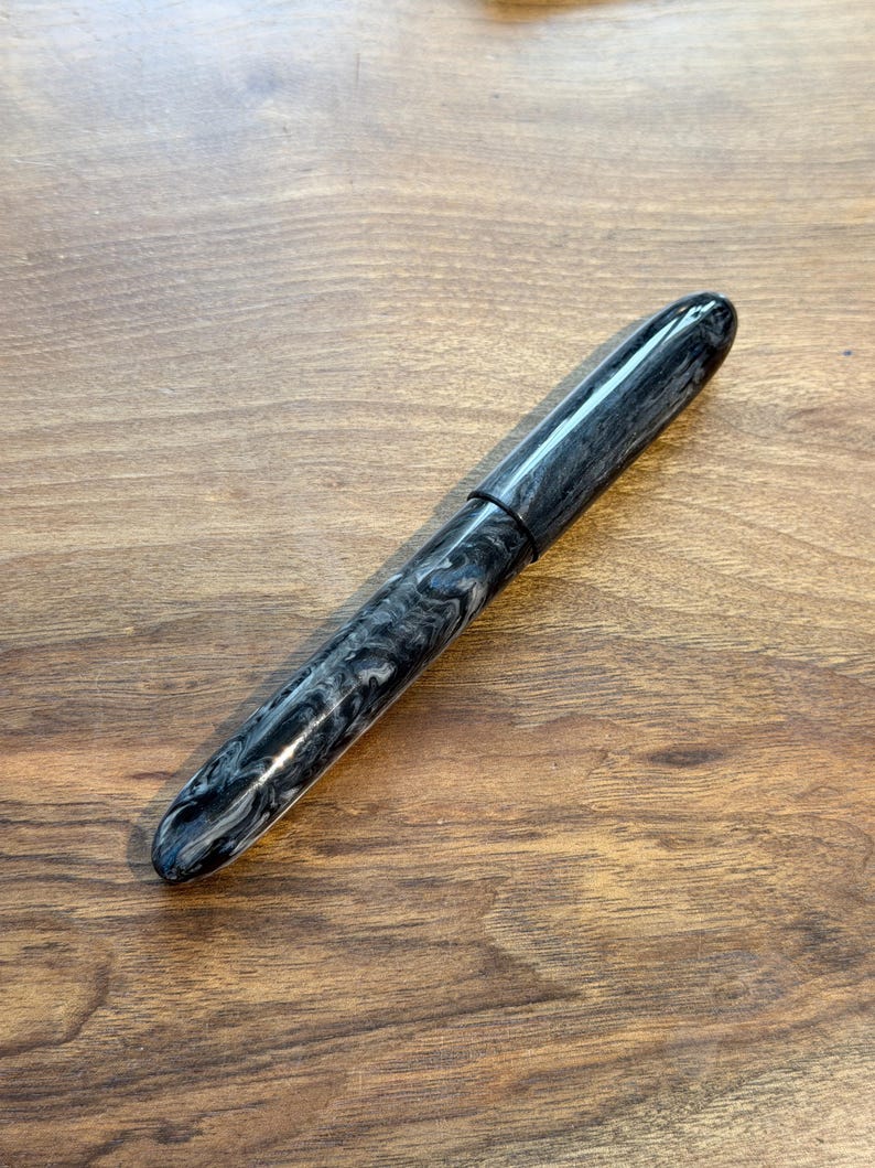 May include: A black and gray marbled fountain pen with a rounded tip and cap. The pen is lying on a wooden surface. The pen's design features a swirling pattern of black and gray, creating a unique aesthetic. The pen is a writing instrument.